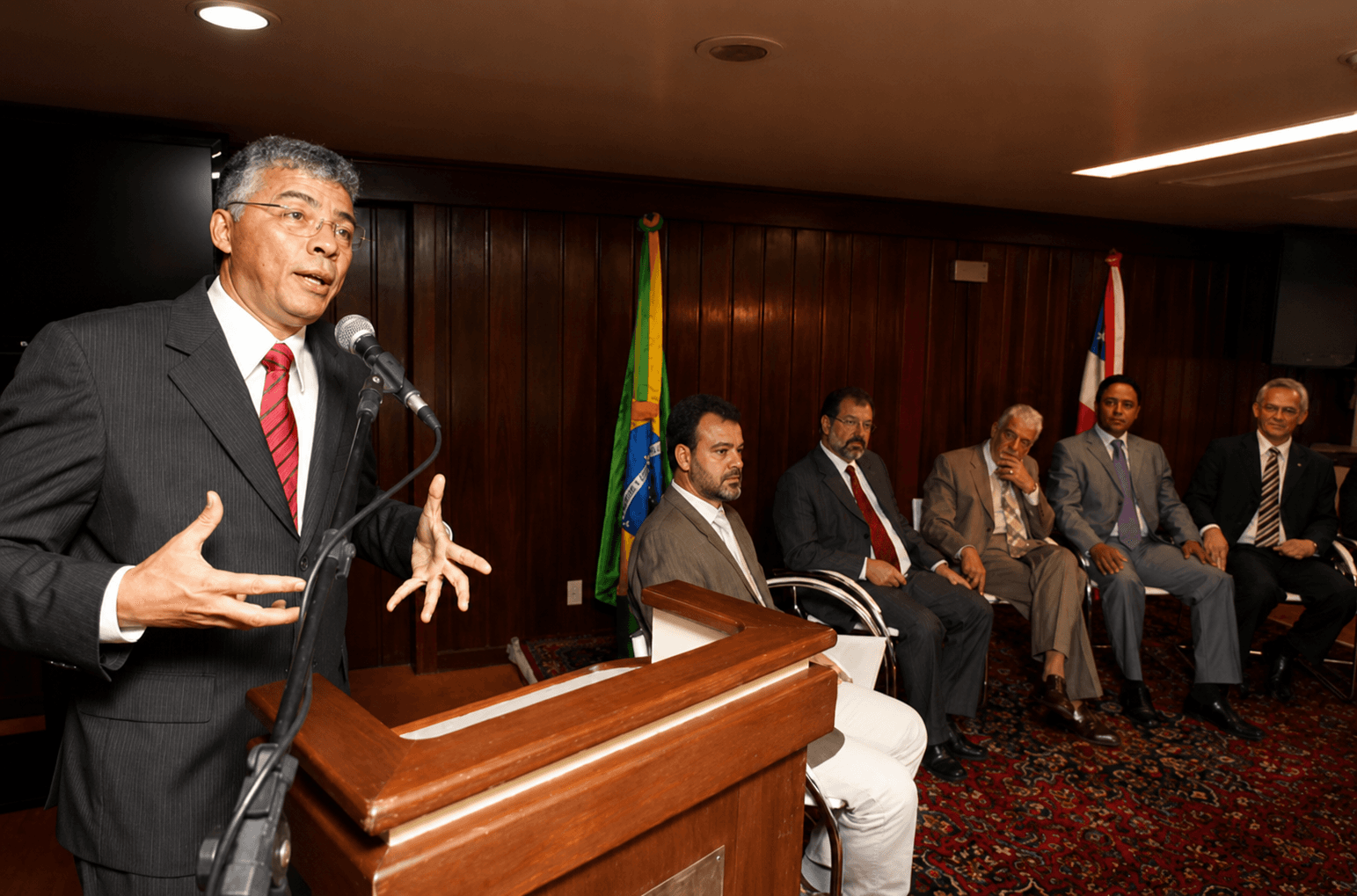 Professor Ney Campello discursando ao púlpito, com as bandeiras do Brasil e da Bahia