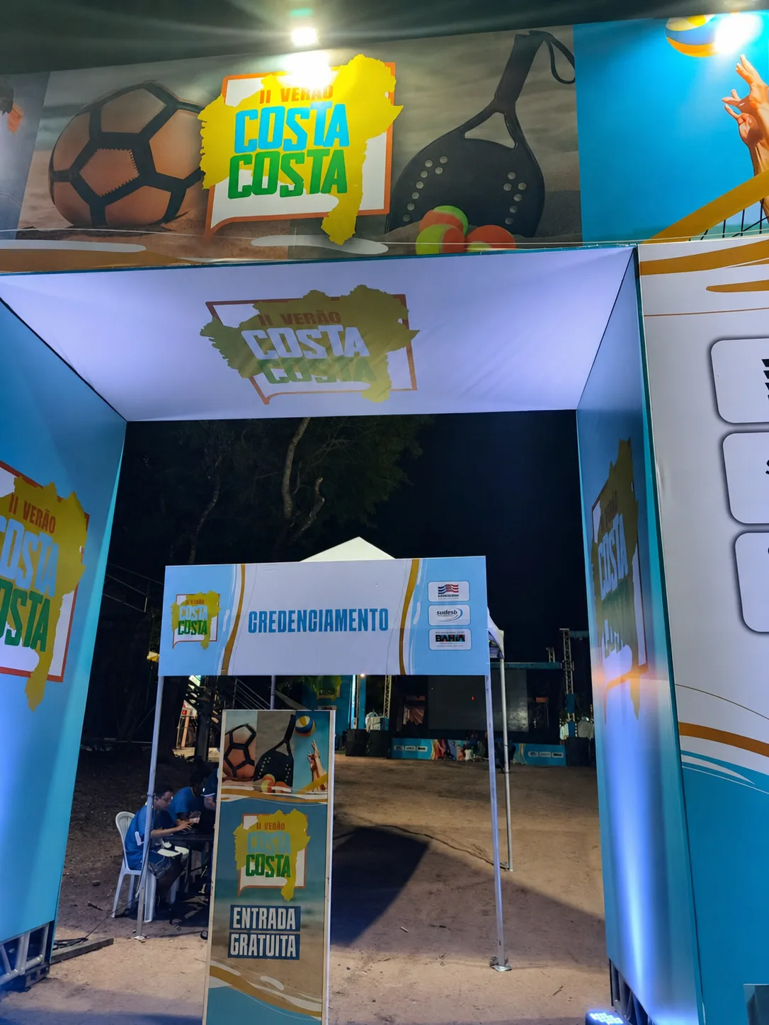 Portal de entrada do II Verão Costa a Costa à noite, com a tenda de credenciamento e a sinalização de entrada gratuita